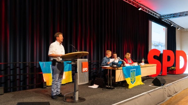 Rainer Albrecht auf der Bühne am Pult bei seiner Vorstellungsrede zur Wahl des Vorsitzenden. Im Hintergrund rechts ist das Präsidium zu sehen sowie große Buchstaben (SPD). Am Präsidiumstisch wird eine Ukraine-Fahne präsentiert.